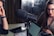 Close-up of a podcast or recording setup featuring a large microphone at the center with two people in the background. One is a woman with glasses, and the other is a person on the left partially out of frame. A laptop and a tablet are on the table.