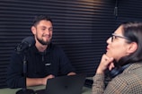 A man and a woman are sitting facing each other in a soundproof room, engaging in a conversation. The man is smiling and seated in front of a microphone, suggesting a podcast or interview setting. The woman is leaning on her hand, wearing glasses and headphones around her neck. A laptop is in front of the man, and the walls are lined with acoustic foam panels.