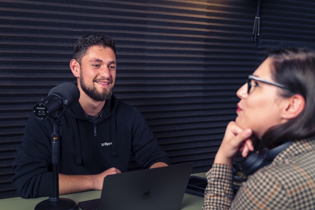 A man and a woman are sitting facing each other in a soundproof room, engaging in a conversation. The man is smiling and seated in front of a microphone, suggesting a podcast or interview setting. The woman is leaning on her hand, wearing glasses and headphones around her neck. A laptop is in front of the man, and the walls are lined with acoustic foam panels.
