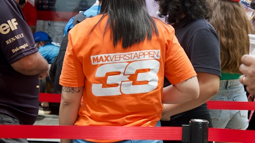 A person is standing with their back to the camera, wearing an orange T-shirt that has the name 'Max Verstappen' and the number '33' printed on it. They are in a crowd, indicated by other people nearby, some wearing shirts with logos. The scene is cordoned off by red ropes.
