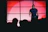 A person stands on a stage, in front of a red and white backdrop consisting of square panels. The individual is wearing a black shirt and appears to be speaking or performing, with an audience visible in silhouette in the foreground.