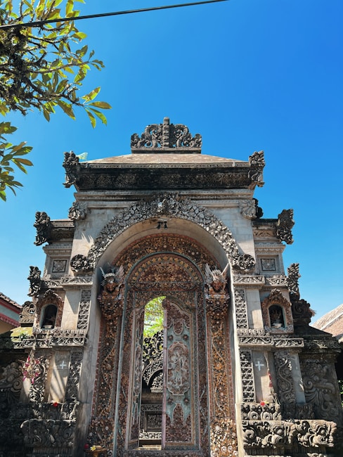 An intricately designed stone gateway exhibits elaborate carvings and detailed artwork. Various decorative elements, including floral and mythical motifs, adorn the structure. The bright blue sky and surrounding greenery complement the historical and cultural architectural style.