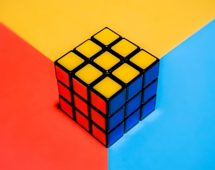 A Rubik's cube is placed on a tri-colored background divided into red, yellow, and blue sections. The cube is solved, with each visible face showing a solid color: yellow, red, and blue.