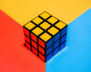 A Rubik's cube is placed on a tri-colored background divided into red, yellow, and blue sections. The cube is solved, with each visible face showing a solid color: yellow, red, and blue.