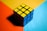 A solved Rubik's Cube is resting on a multi-colored surface. The top face consists of nine yellow squares, while the visible side faces are blue. The background is divided into three sections with bold colors: orange, yellow, and light blue.