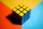 A solved Rubik's Cube is resting on a multi-colored surface. The top face consists of nine yellow squares, while the visible side faces are blue. The background is divided into three sections with bold colors: orange, yellow, and light blue.