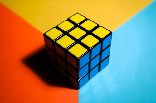 A solved Rubik's Cube is resting on a multi-colored surface. The top face consists of nine yellow squares, while the visible side faces are blue. The background is divided into three sections with bold colors: orange, yellow, and light blue.