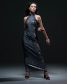 A woman poses confidently in a dark, shimmering, sequined gown. The dress is floor-length with a high neckline and bare arms. She stands against a dark background, with dramatic lighting highlighting the texture and sparkle of the fabric. Her dark hair flows around her face, complementing her bold makeup.