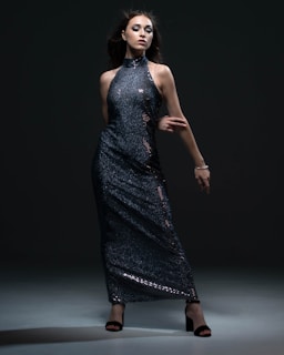A woman poses confidently in a dark, shimmering, sequined gown. The dress is floor-length with a high neckline and bare arms. She stands against a dark background, with dramatic lighting highlighting the texture and sparkle of the fabric. Her dark hair flows around her face, complementing her bold makeup.