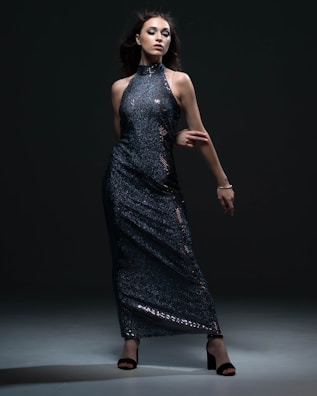 A woman poses confidently in a dark, shimmering, sequined gown. The dress is floor-length with a high neckline and bare arms. She stands against a dark background, with dramatic lighting highlighting the texture and sparkle of the fabric. Her dark hair flows around her face, complementing her bold makeup.