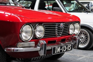 Close-up view of the front end of a classic red car featuring round headlights, a chrome grille, and a license plate with the number IM-13-15. Another vehicle with a different design is partially visible in the background, showcasing its wheel and side panel.