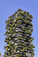 A tall residential building is densely adorned with greenery on each balcony, creating a garden-like appearance against a clear blue sky. The structure features multiple terraces with a variety of trees and shrubs integrated into its design.