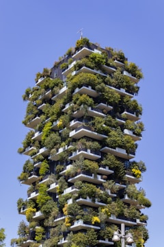 A tall residential building is densely adorned with greenery on each balcony, creating a garden-like appearance against a clear blue sky. The structure features multiple terraces with a variety of trees and shrubs integrated into its design.