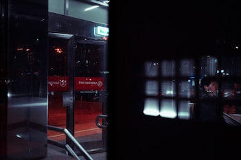 A dimly lit scene featuring an entrance with glass doors and red signs displaying text in Cyrillic. The reflection of a person is visible in the glass, and the interior includes metal railings. The setting appears to be indoors with a view of the street through the doors.