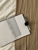 An open planner with a transparent ruler placed diagonally across the pages, resting on a beige textured fabric. A pen with a clip is attached to the notebook.