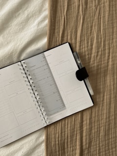 An open planner with a transparent ruler placed diagonally across the pages, resting on a beige textured fabric. A pen with a clip is attached to the notebook.
