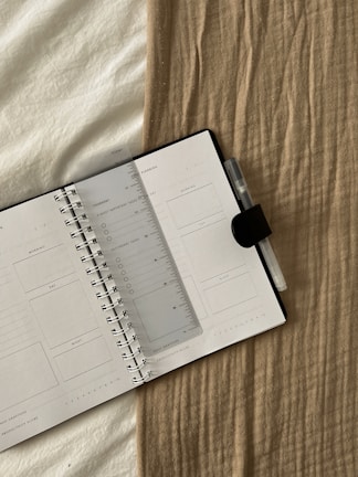 An open planner with a transparent ruler placed diagonally across the pages, resting on a beige textured fabric. A pen with a clip is attached to the notebook.