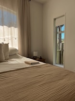 A cozy bedroom bathed in warm sunlight coming through sheer curtains. A neatly made bed takes up most of the frame, with beige bedding and fluffy pillows. On the bedside table sits a small lamp and an open notebook. A narrow window shows a hint of an outdoor scene, possibly a wooden fence and trees.