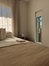 A cozy bedroom bathed in warm sunlight coming through sheer curtains. A neatly made bed takes up most of the frame, with beige bedding and fluffy pillows. On the bedside table sits a small lamp and an open notebook. A narrow window shows a hint of an outdoor scene, possibly a wooden fence and trees.