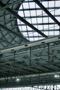 A modern, industrial-style interior roof featuring a large, geometric glass ceiling allowing natural light to illuminate the space. Visible metal beams and support structures complement the grid pattern formed by the glass. Ventilation ducts are prominently displayed, adding to the industrial aesthetic.