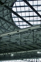 A modern, industrial-style interior roof featuring a large, geometric glass ceiling allowing natural light to illuminate the space. Visible metal beams and support structures complement the grid pattern formed by the glass. Ventilation ducts are prominently displayed, adding to the industrial aesthetic.