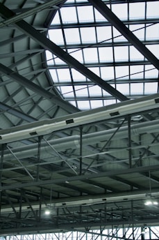 A modern, industrial-style interior roof featuring a large, geometric glass ceiling allowing natural light to illuminate the space. Visible metal beams and support structures complement the grid pattern formed by the glass. Ventilation ducts are prominently displayed, adding to the industrial aesthetic.