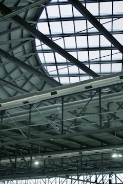 A modern, industrial-style interior roof featuring a large, geometric glass ceiling allowing natural light to illuminate the space. Visible metal beams and support structures complement the grid pattern formed by the glass. Ventilation ducts are prominently displayed, adding to the industrial aesthetic.