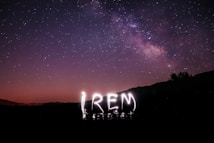 A night sky filled with stars and the Milky Way galaxy visible across the horizon. A word is illuminated in light trails, possibly created with long exposure photography.