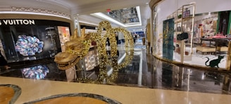 A shopping mall interior with luxury store displays, featuring a prominent golden dragon sculpture adorned with lights. The floor is shiny and reflective, and glass storefronts showcase various items, including high-end handbags and clothing.