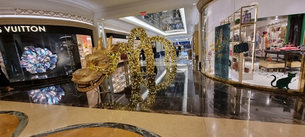 A shopping mall interior with luxury store displays, featuring a prominent golden dragon sculpture adorned with lights. The floor is shiny and reflective, and glass storefronts showcase various items, including high-end handbags and clothing.