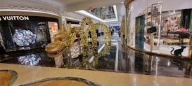 A shopping mall interior with luxury store displays, featuring a prominent golden dragon sculpture adorned with lights. The floor is shiny and reflective, and glass storefronts showcase various items, including high-end handbags and clothing.
