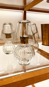 Elegant glass lamps with ribbed designs sit on a wooden shelf with warm lighting in the background. The lamps feature clear glass covers and metallic bases, creating a sophisticated and modern aesthetic. A cylindrical metallic object can be seen in the background, adding to the decorative setup.