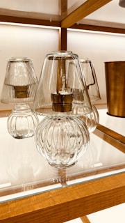 Elegant glass lamps with ribbed designs sit on a wooden shelf with warm lighting in the background. The lamps feature clear glass covers and metallic bases, creating a sophisticated and modern aesthetic. A cylindrical metallic object can be seen in the background, adding to the decorative setup.