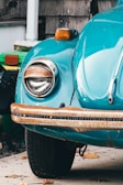 A close-up view of the front left section of an antique car, featuring a teal-colored body and a rusted chrome bumper. The headlight is rounded and the signal light above it glows orange. The surrounding area includes some green bins and a white column, with dried leaves scattered on the ground.