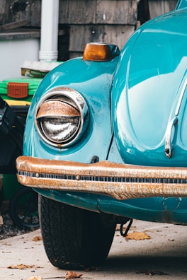 A close-up view of the front left section of an antique car, featuring a teal-colored body and a rusted chrome bumper. The headlight is rounded and the signal light above it glows orange. The surrounding area includes some green bins and a white column, with dried leaves scattered on the ground.