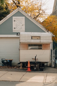 A small trailer is parked in front of a garage with a 'No Parking' sign on the door. The garage has blue-gray siding and white trim, while the trailer appears to be weathered and somewhat old. An orange traffic cone is placed in front of the trailer, and a tree with autumn leaves is visible in the background.