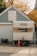 A small trailer is parked in front of a garage with a 'No Parking' sign on the door. The garage has blue-gray siding and white trim, while the trailer appears to be weathered and somewhat old. An orange traffic cone is placed in front of the trailer, and a tree with autumn leaves is visible in the background.