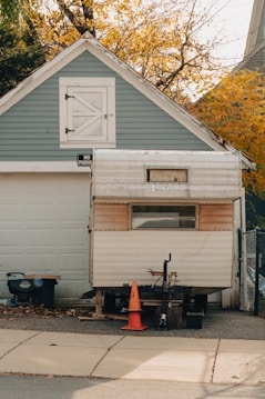 A small trailer is parked in front of a garage with a 'No Parking' sign on the door. The garage has blue-gray siding and white trim, while the trailer appears to be weathered and somewhat old. An orange traffic cone is placed in front of the trailer, and a tree with autumn leaves is visible in the background.