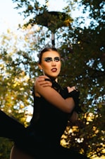 A performer dressed in a dramatic black costume with striking makeup, featuring dark eyeshadow and red lipstick, is standing outdoors. The scene is set against a backdrop of trees with sunlight filtering through the leaves, creating a play of light and shadow.
