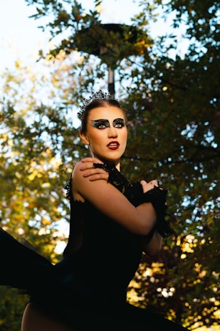 A performer dressed in a dramatic black costume with striking makeup, featuring dark eyeshadow and red lipstick, is standing outdoors. The scene is set against a backdrop of trees with sunlight filtering through the leaves, creating a play of light and shadow.