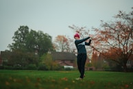 A person is playing golf on a grassy field surrounded by autumn foliage and houses in the background. The golf club is in mid-swing, and the individual is dressed in a blue outfit with a red hat. The scene conveys a calm, overcast day in a suburban setting.