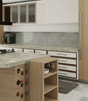 A modern kitchen interior featuring wooden cabinetry and a marble countertop. The kitchen includes a stovetop, a coffee machine, a wine rack with bottles, and open shelving holding wine glasses and a bowl. The backsplash is tiled, and a small rug is placed on the floor.