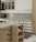 A modern kitchen interior featuring wooden cabinetry and a marble countertop. The kitchen includes a stovetop, a coffee machine, a wine rack with bottles, and open shelving holding wine glasses and a bowl. The backsplash is tiled, and a small rug is placed on the floor.