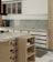 A modern kitchen interior featuring wooden cabinetry and a marble countertop. The kitchen includes a stovetop, a coffee machine, a wine rack with bottles, and open shelving holding wine glasses and a bowl. The backsplash is tiled, and a small rug is placed on the floor.