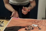 A person with tattoos and a bracelet is holding a red pen over a colorful illustration. The illustration includes abstract designs and vibrant colors, with a pink sticky note labeled 'Sold' attached to it.