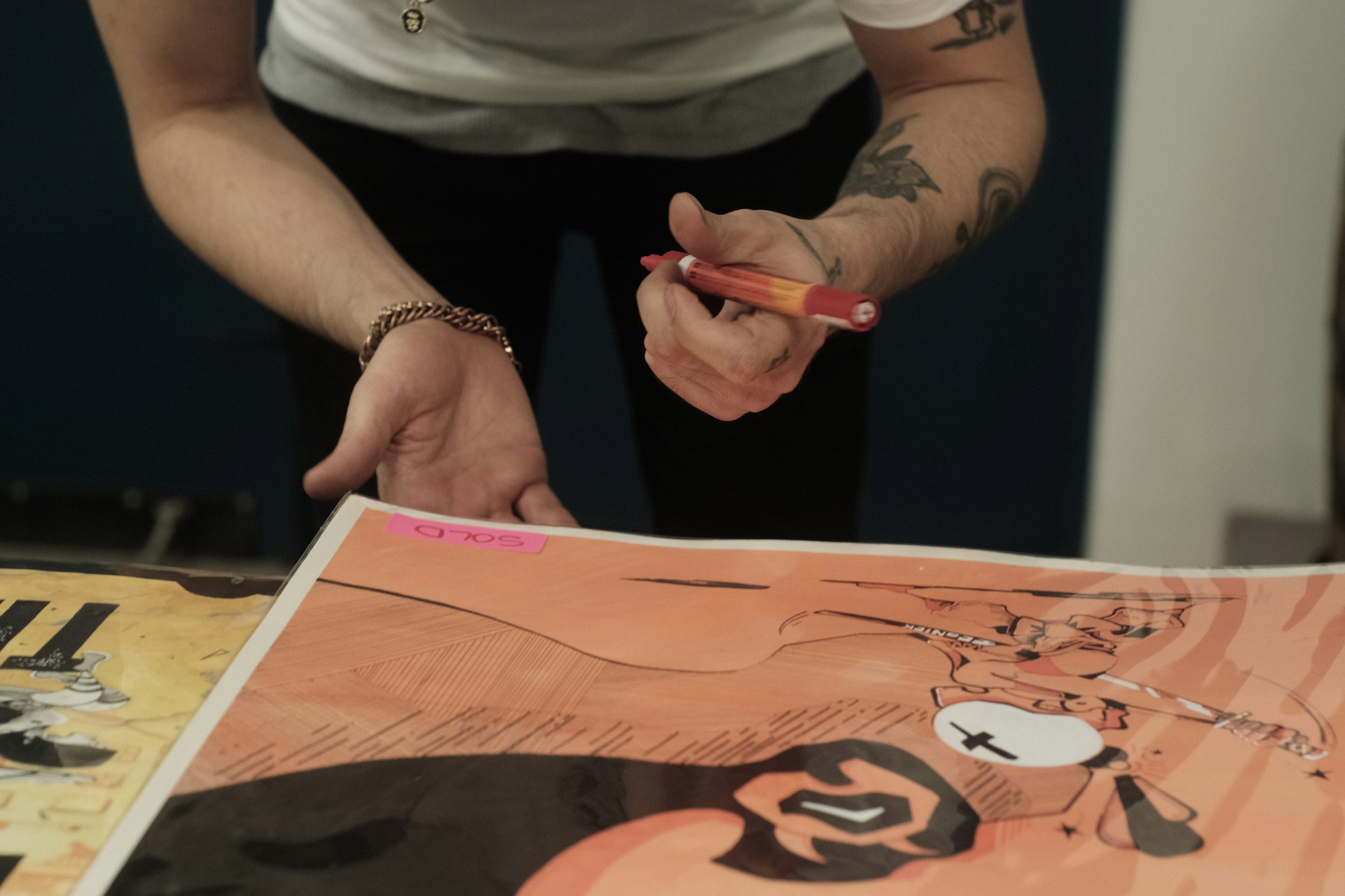 A person with tattoos and a bracelet is holding a red pen over a colorful illustration. The illustration includes abstract designs and vibrant colors, with a pink sticky note labeled 'Sold' attached to it.