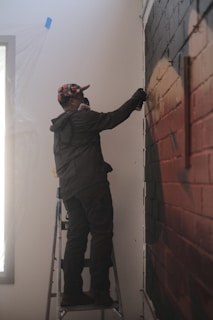 A person standing on a ladder is spray painting a mural on a brick wall. The individual is wearing a colorful cap and a dark jacket, with a protective mask covering their face. The surrounding area appears to be a room with some plastic covering near a window.