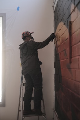 A person standing on a ladder is spray painting a mural on a brick wall. The individual is wearing a colorful cap and a dark jacket, with a protective mask covering their face. The surrounding area appears to be a room with some plastic covering near a window.