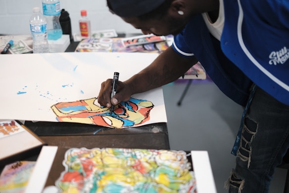 A person is drawing on a large sheet of paper using vibrant colors. The art appears to be expressive and abstract, featuring bold strokes and splashes of blue, red, and yellow. Nearby, assorted drawing tools and bottles, including water bottles and spray paint, are scattered on the table.