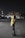 A person stands outdoors at night on an empty street, wearing a yellow sports jersey over a hoodie. The background consists of dimly lit buildings and streetlights, with a car parked on one side.